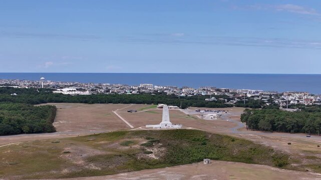 An expansive aerial shot shows a circular road loop around a grassy hill topped by a White Wright Brothers monument, with distant Kill Devil Hills and Outer Banks Coastline under a clear blue sky.