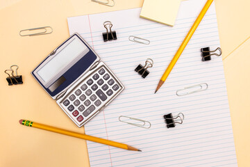 horizontal flat lay of office supplies: handheld calculator, paper clips, pencils, manila folders, lined paper, and binder clips