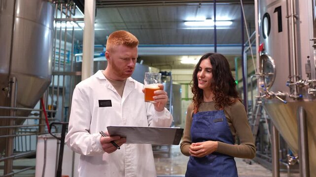 Male food technologist in a lab coat performing quality control on a beer sample with a female brewer inside a craft beer factory