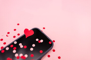 close-up of a cellphone with a black screen with pink and red confetti and a cutout paper heart against a pink background with copy space available