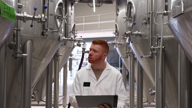 Young male brewery technologist in a white lab coat walking through rows of large steel vats, inspecting equipment and taking notes