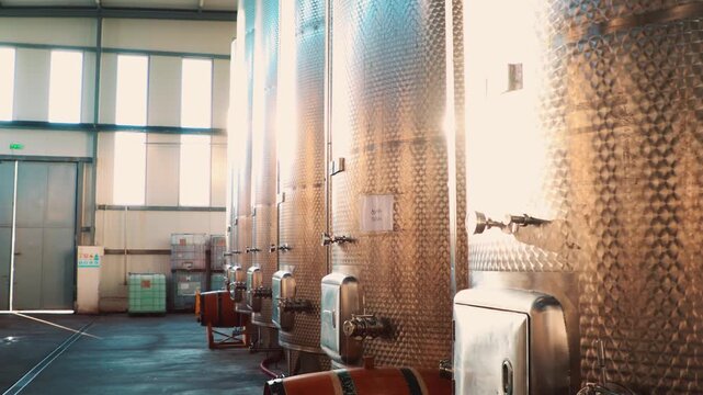 Large industrial metal wine tanks with valves, pressure gauges, and fittings lined up in a row inside a modern winery production facility showcasing stainless steel fermentation vessels. Pan shot.