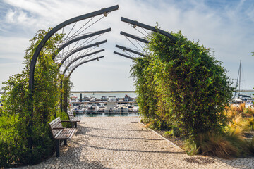 Wooden benches sit under a vine-covered metal archway overlooking boats docked at the marina.
