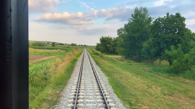 A view from the window of the last carriage of a passing train onto a railway running through a rural area in the rays of the setting sun in Poland.