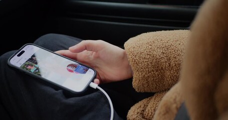 Close-up of a hand in a fleece jacket using a smartphone with a charging cable inside a car. The screen shows a social media app, representing mobile browsing, communication, and relaxed lifestyle. - Powered by Adobe