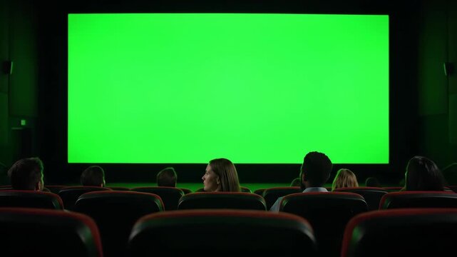 Audience in a movie theater watching a large green screen. People sitting in a dark cinema looking at a chroma key display. Mockup with copy space for advertising or film presentation