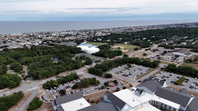 An expansive aerial shot shows a circular road loop around a grassy hill topped by a White Wright Brothers monument, with distant Kill Devil Hills and Outer Banks Coastline under a clear blue sky.