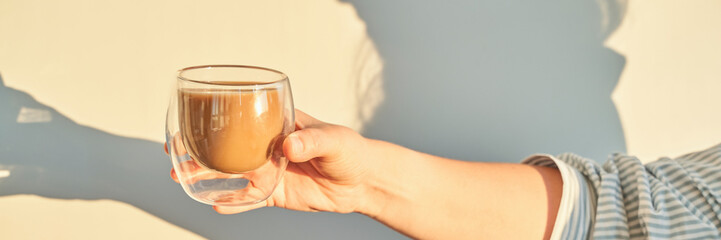 Caucasian adult holding glass cup with coffee reflecting warm light and shadow.