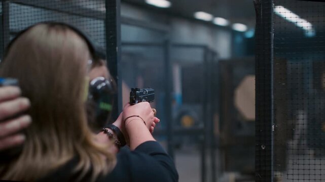 Woman with gun shoots a firearm at a shooting gallery or range. Female is engaged in sports shooting, wearing headphones. Pistol is a small weapon. Cartridge case goes off in slow motion