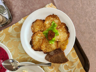 plate of golden-brown potato pancakes (likely Draniki or Latkes) served with a dollop of topping, lettuce, and a slice of dark bread, partially obscuring a bowl of red soup.
