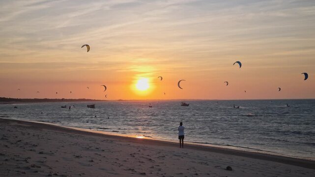 Beach sunset at Barra Grande, Piau&iacute;. Kitesurfers and boats on blue sea, person on sandy shore, vivid orange sky. Tropical adventure and peaceful vacation scenery, Brazil.