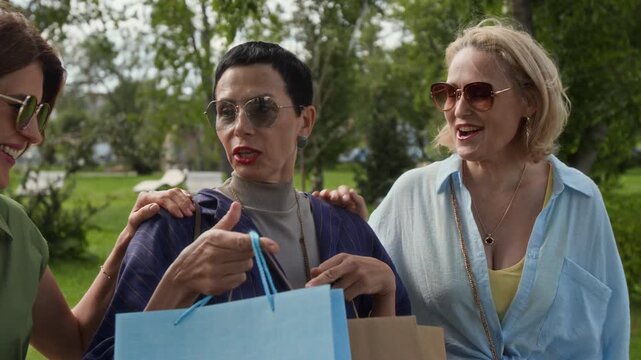 Three joyful middle-aged Caucasian friends looking at goods in shopping bags while walking outdoors on summer day