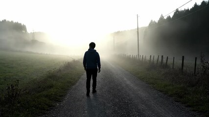A man walks alone on a gravel road shrouded in early morning fog and sunlight. - Powered by Adobe