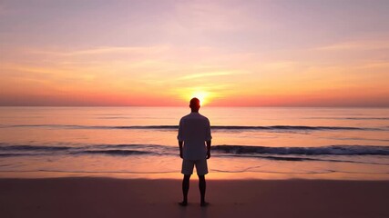 Man stands on beach, facing ocean at sunrise, pink and orange sky - Powered by Adobe