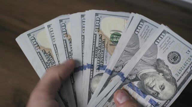 Top view of male hands holding and inspecting a single one hundred dollar bill over a dark wood table, checking the US currency for authenticity by showing the front and back of the banknote.