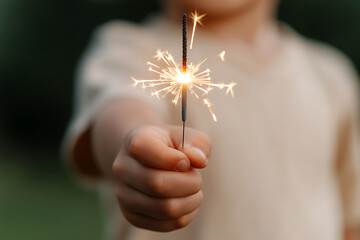 A child's hand holds a sparking sparkler, illuminating the frame with bursts of light. Celebratory glow in a tender moment. Simple joys of childhood.