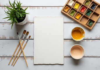 Top view of a watercolor painting setup with brushes, paints, paper, and a plant on a wooden table
