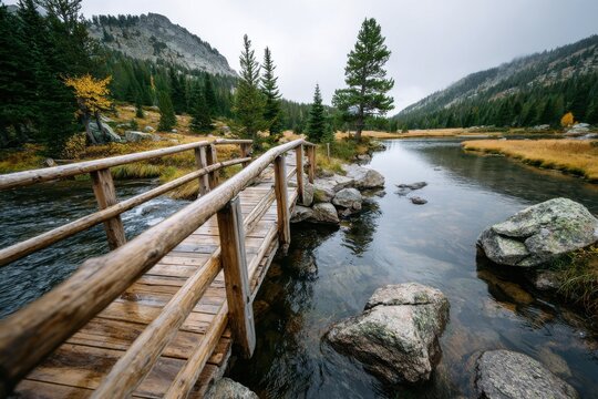 Scenic wooden bridge over a calm river surrounded by tall trees and mountains on a cloudy autumn day