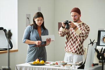 Man photographing food on table with digital camera while woman holding tablet standing beside him, both focused on food styling process