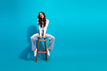 Happy young woman sitting on a wooden stool with a bright casual outfit against a seamless vibrant blue background