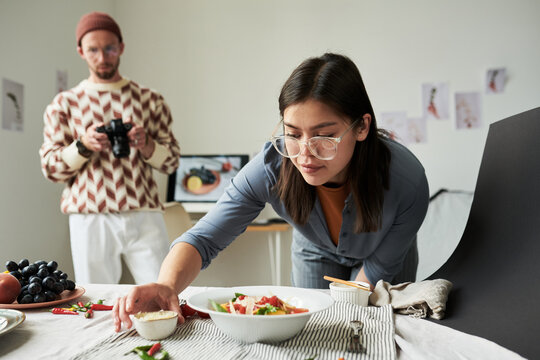 Asian woman arranging food on table while man with beard photographing scene in background, creative workspace with food styling setup visible