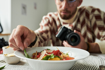 Man holding camera arranging garnish on salad in bowl, focusing on food styling for photography, wearing glasses and patterned sweater, sitting at table