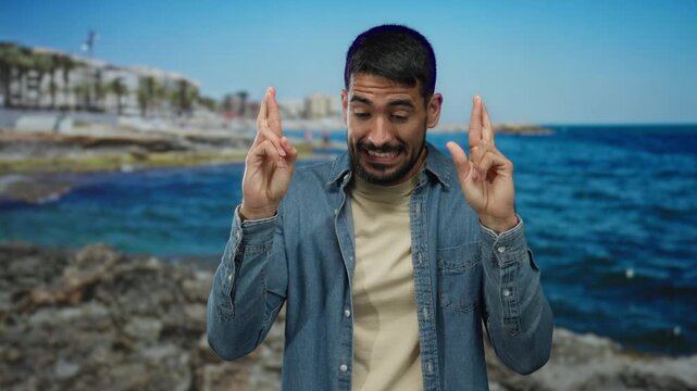 Young man crossing fingers with a hopeful expression at the seaside, wearing casual denim attire on a sunny beach day, reflecting optimism and anticipation outdoors.