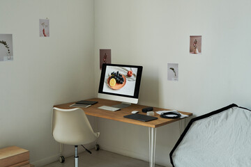 Minimalist workspace featuring desktop computer displaying fruit plate image, wireless keyboard, mouse, tablet, smartphone, headphones on wooden desk against wall with small art prints