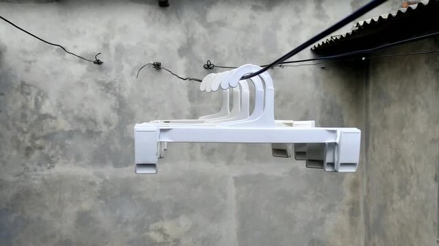Five white plastic hangers hang on a clothesline on a textured gray wall, with a cable stretching across the top of the frame. The hangers sway in the wind.