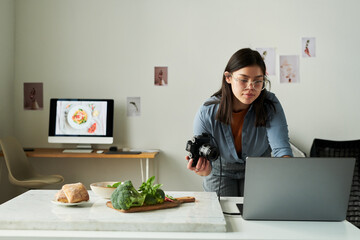 Woman wearing glasses holding digital camera while working on laptop in modern kitchen, preparing food photography setup with vegetables and bread on counter
