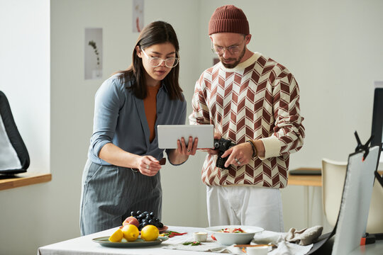 Asian woman and man collaborating over digital tablet and camera, preparing food photography setup on table with fruits and plates in modern workspace - Powered by Adobe