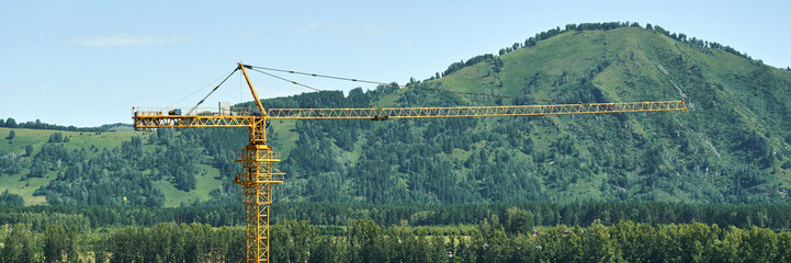 Large yellow construction crane against forested mountain backdrop under clear blue sky.