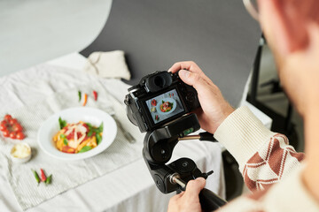 Man photographing plate of food with professional camera, capturing overhead shot of colorful meal on table, hands adjusting camera settings during food photography session