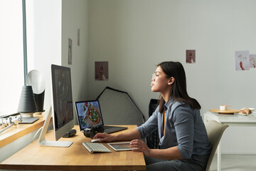 Asian woman working at desk using desktop computer and digital tablet, analyzing food images on screen, camera and laptop nearby, sitting in modern workspace