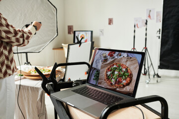Woman photographing pizza with smartphone while laptop displaying food image on screen, professional food photography studio setup with lighting equipment visible