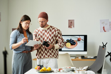Man holding camera standing next to Asian woman writing on digital tablet, both collaborating in modern photography studio with food on table