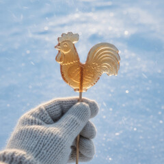 Fototapeta premium Knitted glove holding a caramel rooster lollipop in gentle snowfall with warm festive bokeh on a cold blue background. Cozy winter holiday mood with copy space.