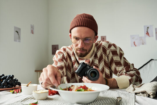 Man wearing glasses holding camera styling food on table preparing for food photography session focusing on arranging ingredients in bowl - Powered by Adobe
