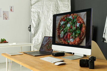 Modern workspace featuring desktop computer and laptop displaying close up of gourmet pizza with fresh toppings, digital camera and tablet resting on wooden desk in photography studio