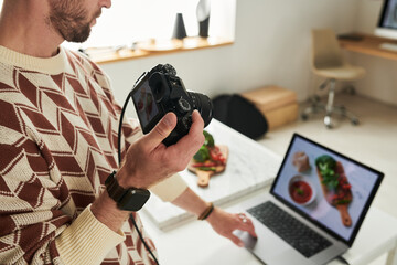Man holding digital camera while reviewing food photographs on laptop, standing at table with...