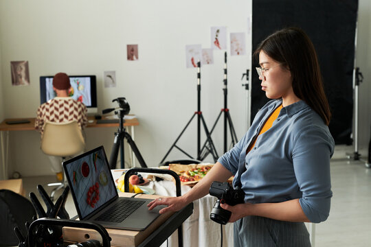 Asian woman holding digital camera reviewing food photographs on laptop in professional photo studio