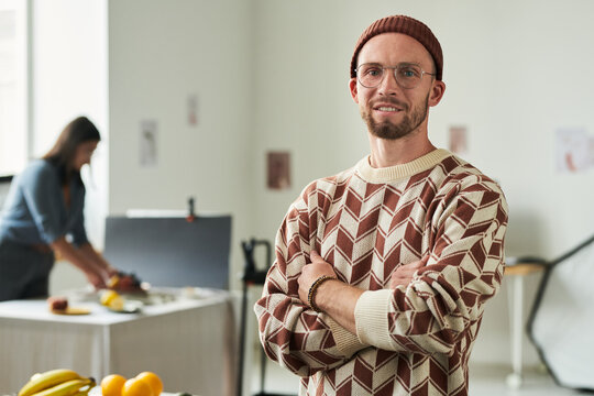 Portrait of man with beard and glasses standing with arms crossed in bright studio, woman working at table in background, creative workspace setting