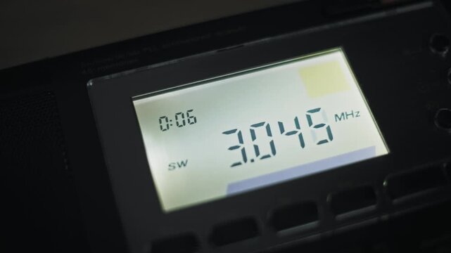 Angled macro of a digital radio tuner scanning up the shortwave band showing changing MHz display signal strength meter and PLL synthesized receiver text on a black device in low light.