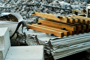 A dynamic horizontal shot of various building materials on a construction site. Features a pile of granite cobblestones, stacked metal roofing sheets, timber planks, and concrete blocks.