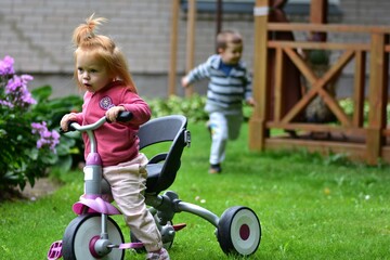 Red-haired toddler girl riding a tricycle on green grass while a boy runs behind her, both enjoying playful outdoor moments and active fun in the backyard garden.