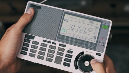 A person holds and tunes a portable multi-band digital radio. Fingers press buttons to switch between FM LW MW and SW bands with the LCD screen displaying corresponding frequency and band data.