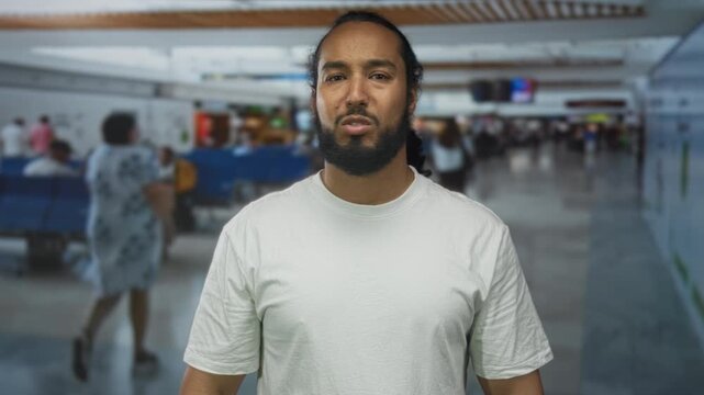 Man with beard and clasped hands at busy airport terminal in white t shirt, standing amid seating and travelers, facing camera; concern travel.