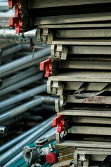 A vertical shot displays neatly stacked aluminum scaffolding components with red latches, alongside a blurred background of metal pipes. This illustrates organized construction equipment storage.