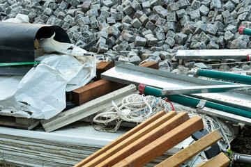 A cluttered view of a construction site showing mixed materials: granite cobblestones, rolled waterproofing membrane, wood planks, metal profiles, and tangled ropes, representing site cleanup.