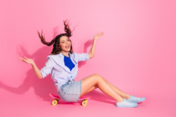 Pretty woman sitting on a skateboard on a pink background with a cheerful expression and playful...